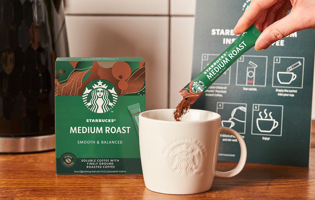 一包星巴克優質即溶中度烘焙咖啡倒入印有 We Proudly Serve Starbucks® 星巴克咖啡服務字樣的陶瓷馬克杯中