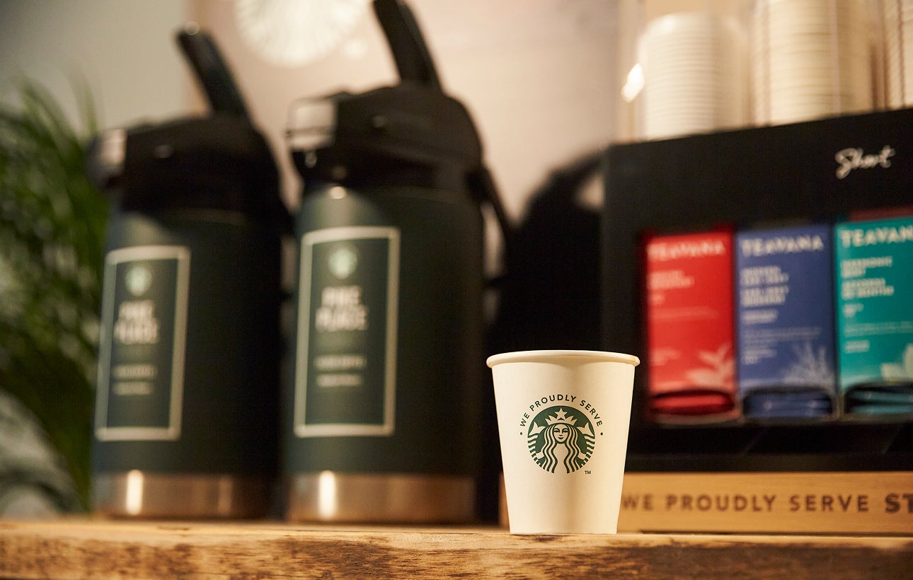 一個印有 We Proudly Serve Starbucks 星巴克咖啡服務字樣的紙杯放在大型現煮咖啡機前的木製櫃檯上