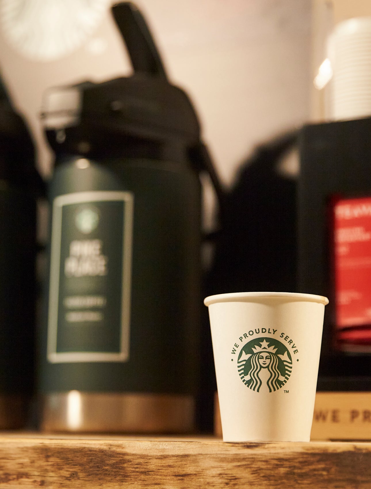 一個印有 We Proudly Serve Starbucks 星巴克咖啡服務字樣的紙杯放在大型現煮咖啡機前的木製櫃檯上