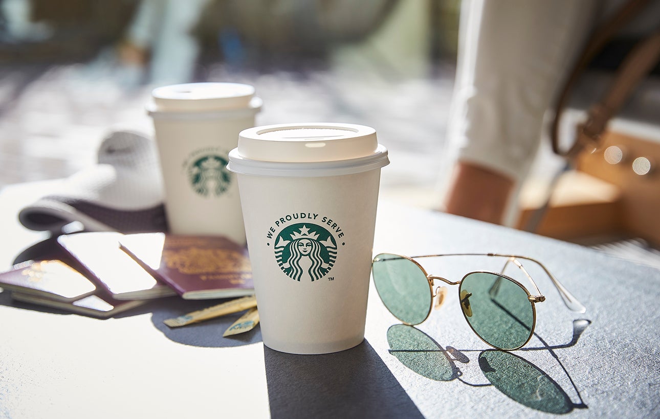 兩個印有 We Proudly Serve Starbucks 星巴克咖啡服務字樣的外帶杯 (有蓋子) 擺放在戶外桌上，旁邊還有一副太陽眼鏡