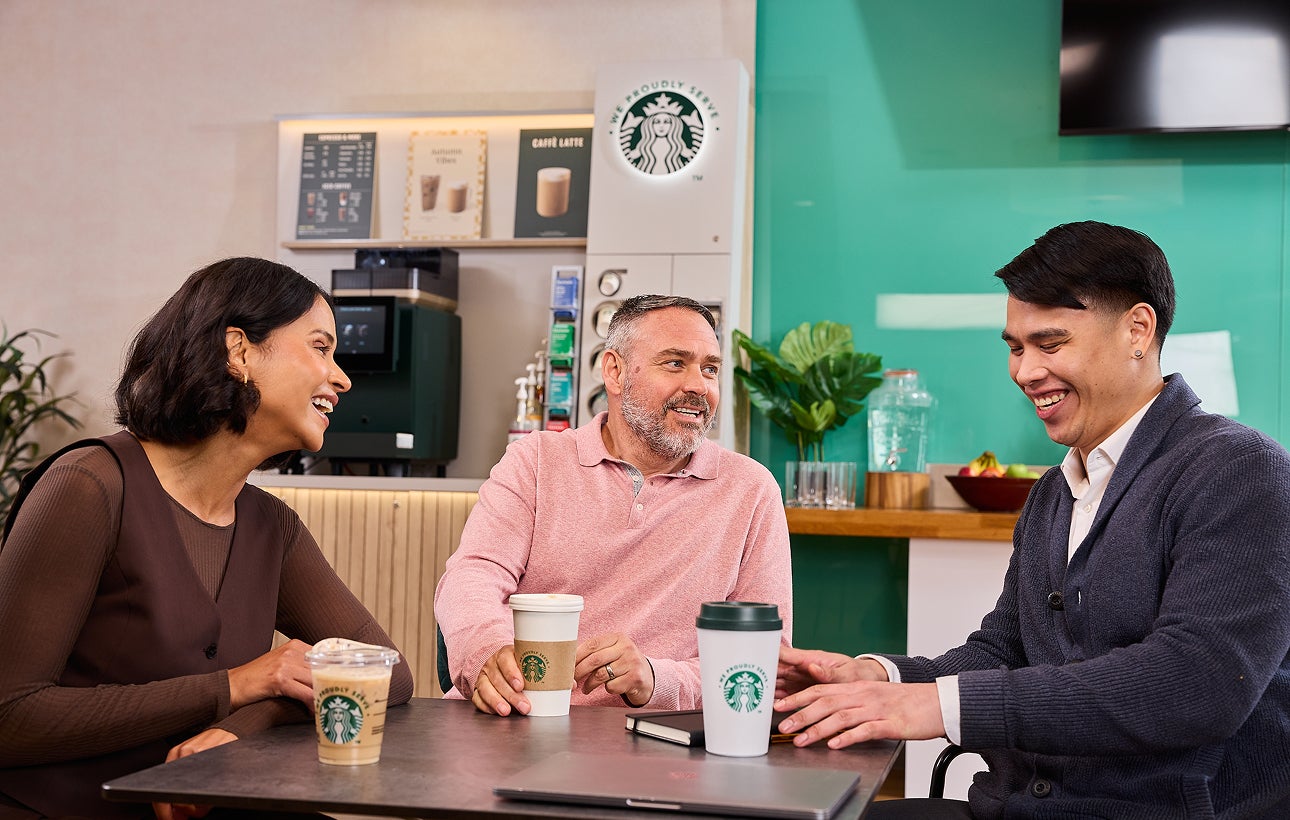同事們坐在一起，背景中可以看到印有 We Proudly Serve Starbucks 星巴克咖啡服務字樣的杯子和自助咖啡機