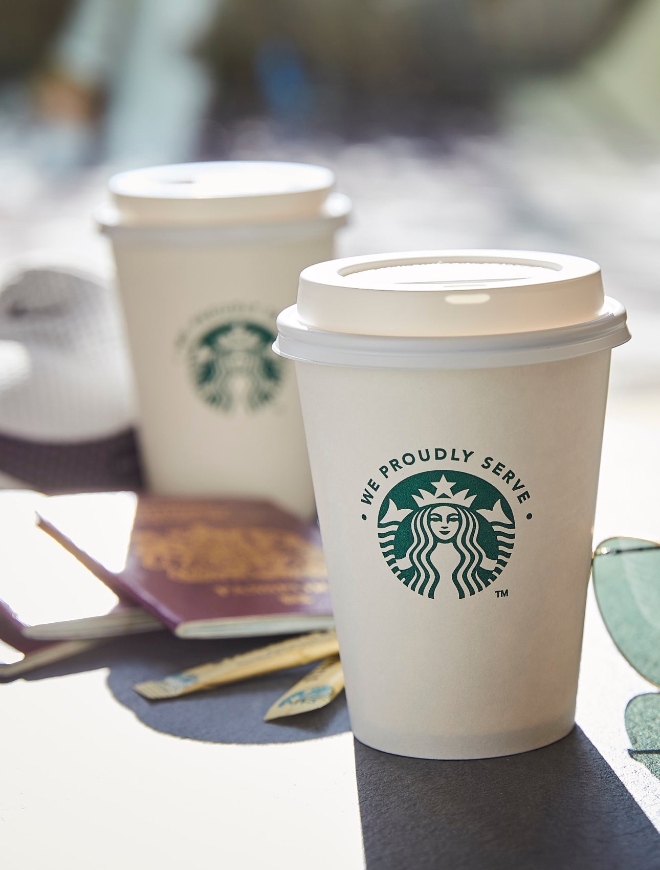 兩個印有 We Proudly Serve Starbucks 星巴克咖啡服務字樣的外帶杯 (有蓋子) 擺放在戶外桌上,旁邊還有一副太陽眼鏡