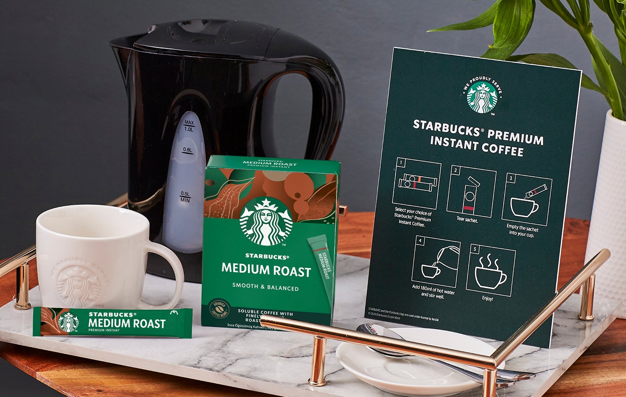 桌面上擺著印有 We Proudly Serve Starbucks 星巴克咖啡服務字樣的馬克杯、星巴克即溶咖啡和沖泡說明