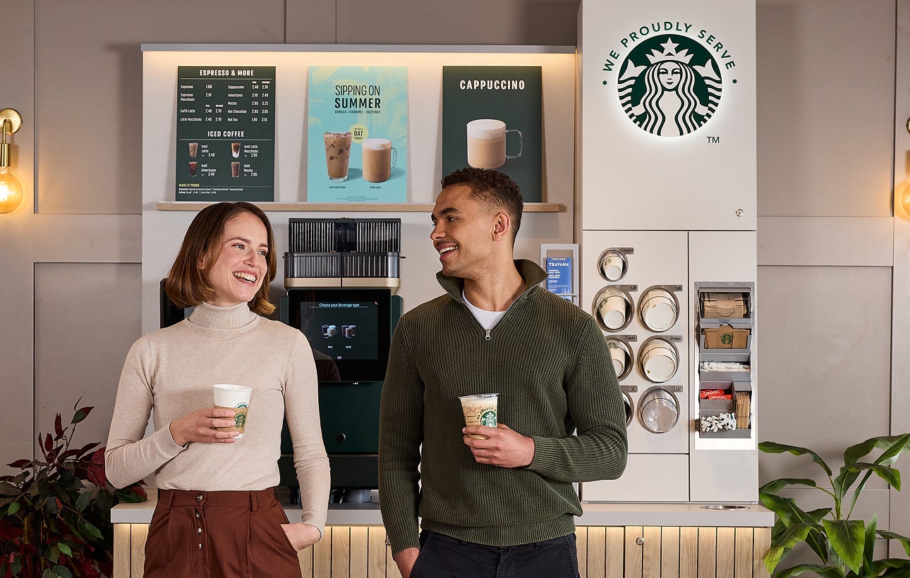 兩個人站在自助咖啡機前，一邊聊天一邊拿著印有 We Proudly Serve Starbucks 星巴克咖啡服務字樣的咖啡杯