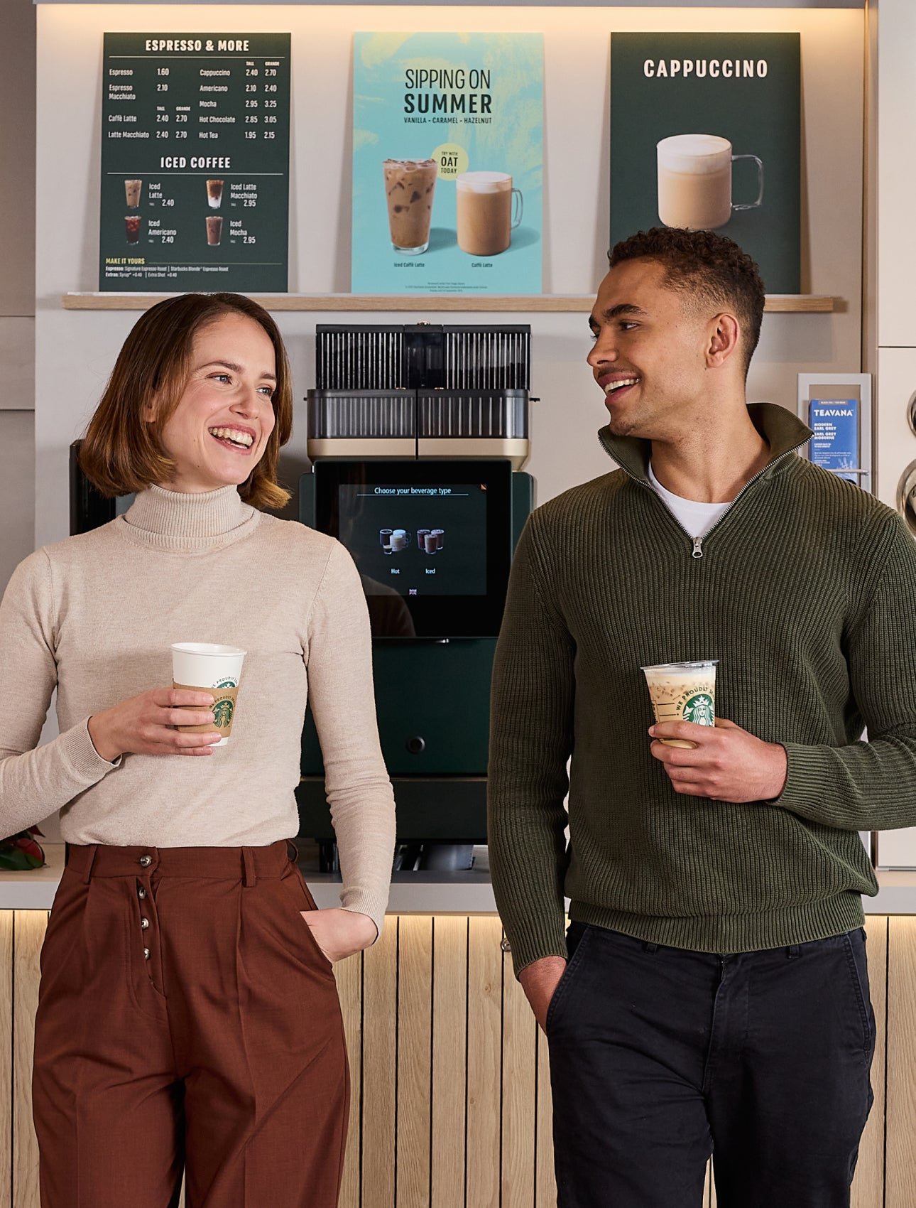 兩個人站在自助咖啡機前，一邊聊天一邊拿著印有 We Proudly Serve Starbucks 星巴克咖啡服務字樣的咖啡杯