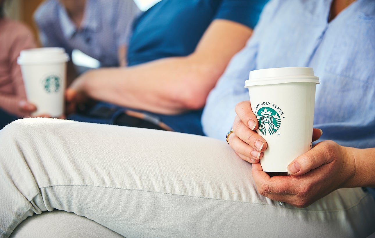 一個人坐著，雙手拿著一個印有 We Proudly Serve Starbucks 星巴克咖啡服務字樣的外帶杯