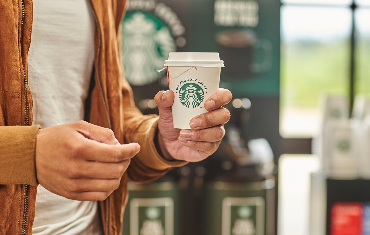 一個人手裡拿著一杯茶，杯子上印著 We Proudly Serve Starbucks 星巴克咖啡服務字樣