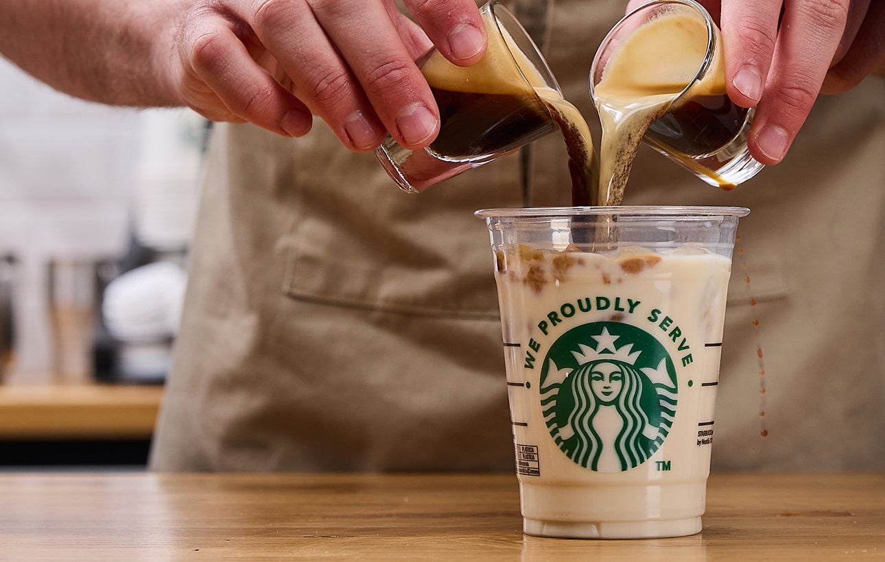 咖啡師同時將兩份濃縮咖啡倒入印有 We Proudly Serve Starbucks 星巴克咖啡服務字樣的杯子中，杯子裡裝了冰塊和牛奶