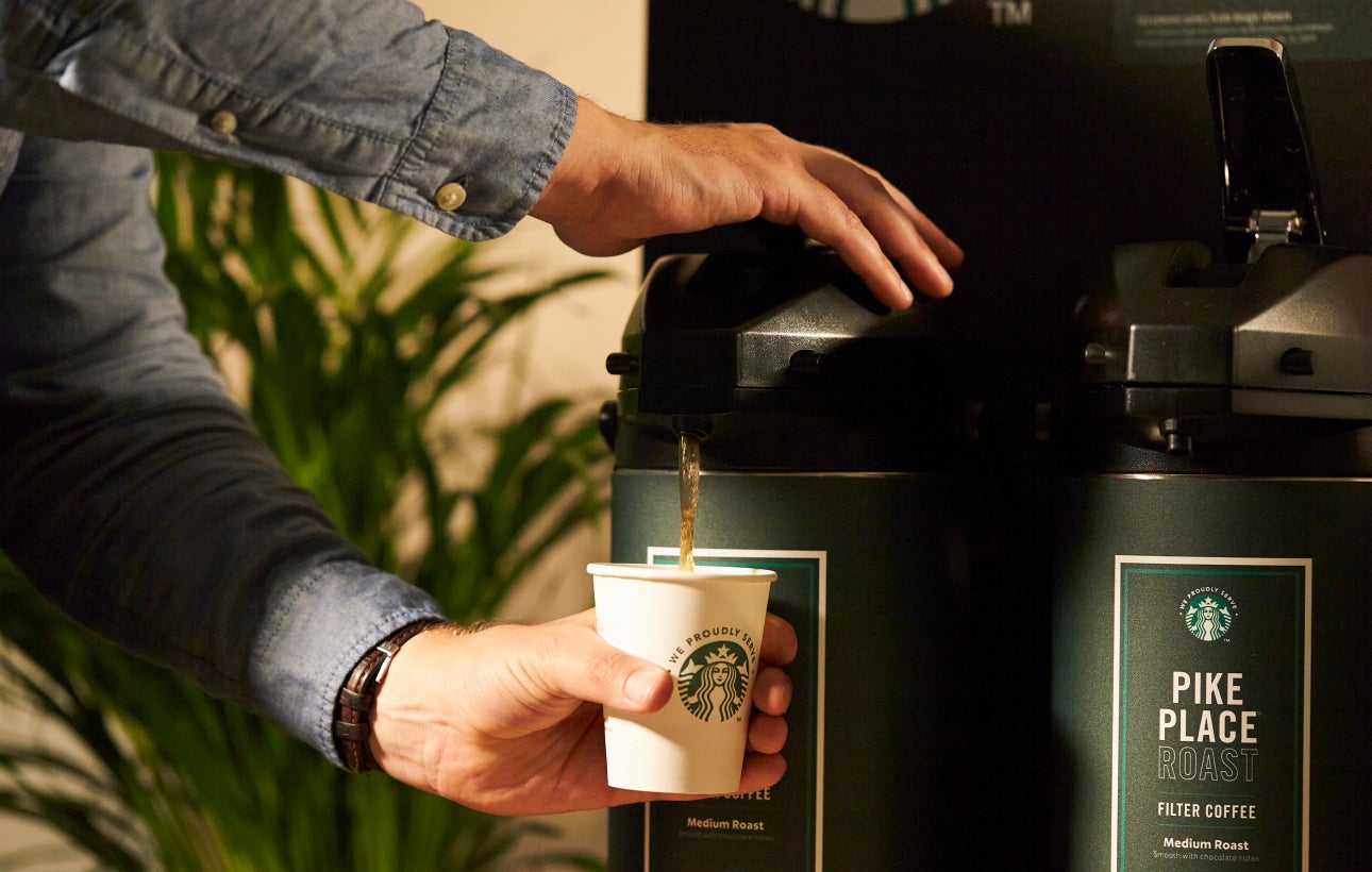 一個人從大型咖啡機將星巴克 Pike Place™ 派克市場烘焙咖啡倒入印有 We Proudly Serve Starbucks® 星巴克咖啡服務字樣的杯子中