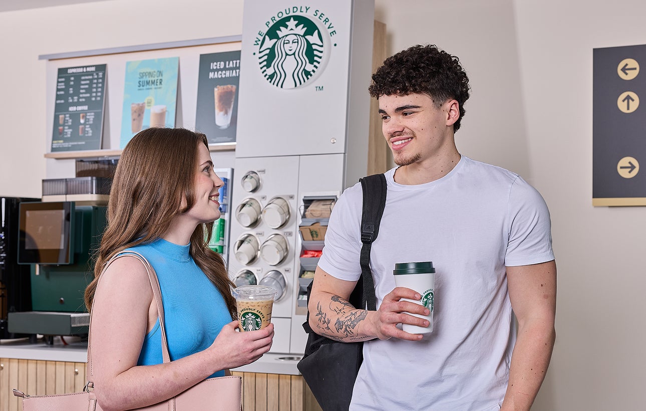 一男一女站在 We Proudly Serve Starbucks 星巴克咖啡服務自助咖啡站前