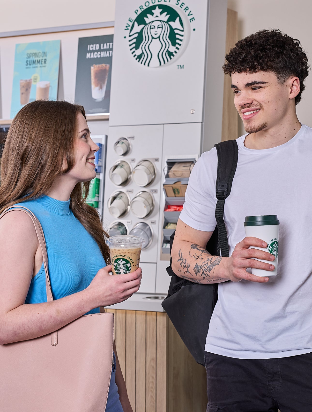一男一女站在 We Proudly Serve Starbucks 星巴克咖啡服務自助咖啡站前