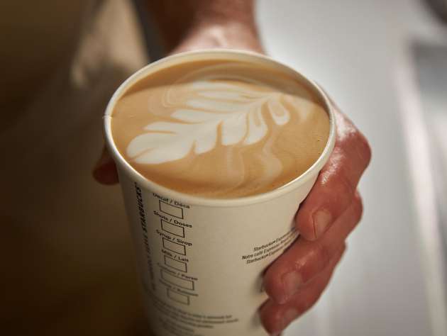 一手拿著一杯印有 We Proudly Serve Starbucks 星巴克咖啡服務字樣的咖啡，上面還有拉花圖案
