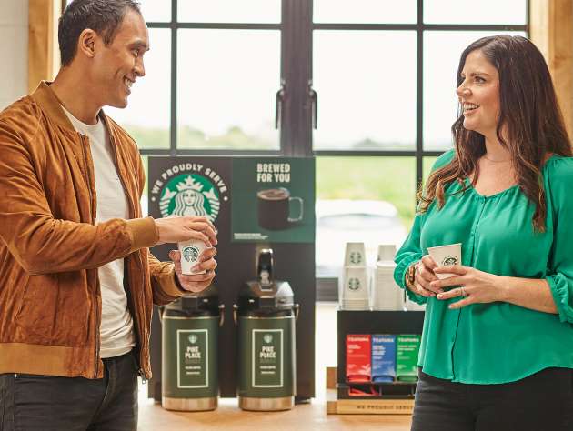 兩個人站在現煮咖啡機前，手持印有 We Proudly Serve Starbucks 星巴克咖啡服務字樣的杯子，站著聊天