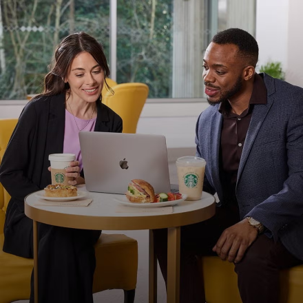 兩個人坐在筆記型電腦前，桌上擺著 We Proudly Serve Starbucks 星巴克咖啡服務咖啡和三明治