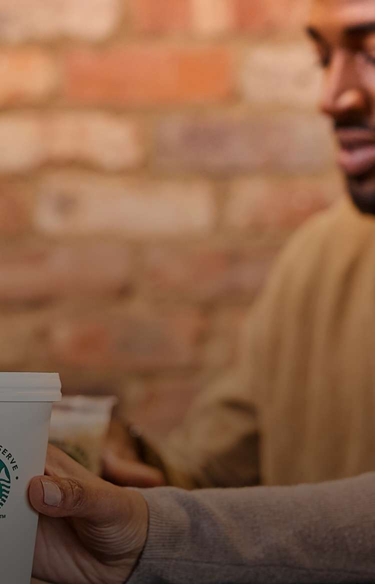 一個人手持印有 We Proudly Serve Starbucks 星巴克咖啡服務字樣的杯子，背景是模糊的磚牆