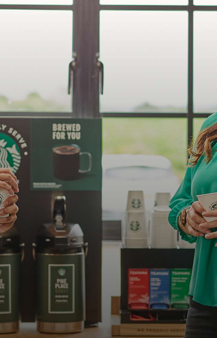 兩個人站在現煮咖啡機前，手持印有 We Proudly Serve Starbucks 星巴克咖啡服務字樣的杯子，站著聊天
