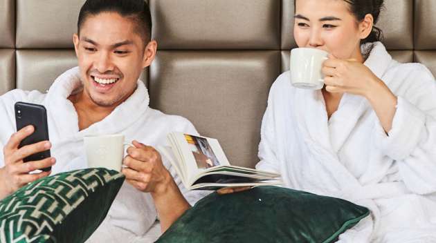 一對情侶穿著白色浴袍，在飯店床上放鬆身心，享用星巴克咖啡。 